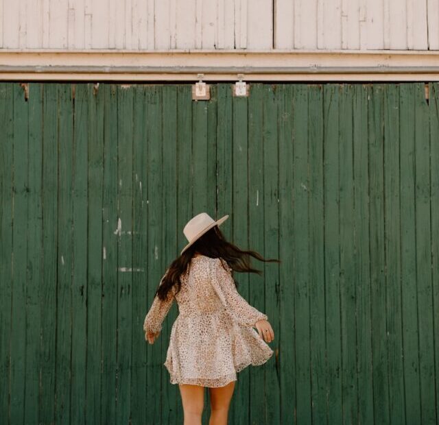 woman in brown and white polka dot dress wearing black hat standing in front of green